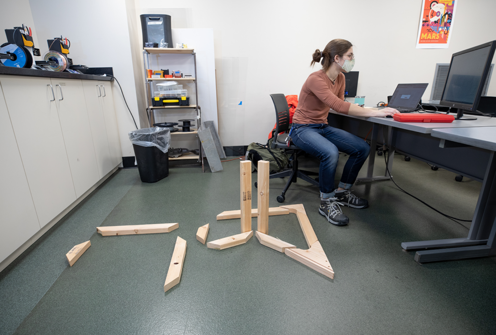 Bridgette Dalldorf sits at the computer with wood for the chair project she is building next to her.