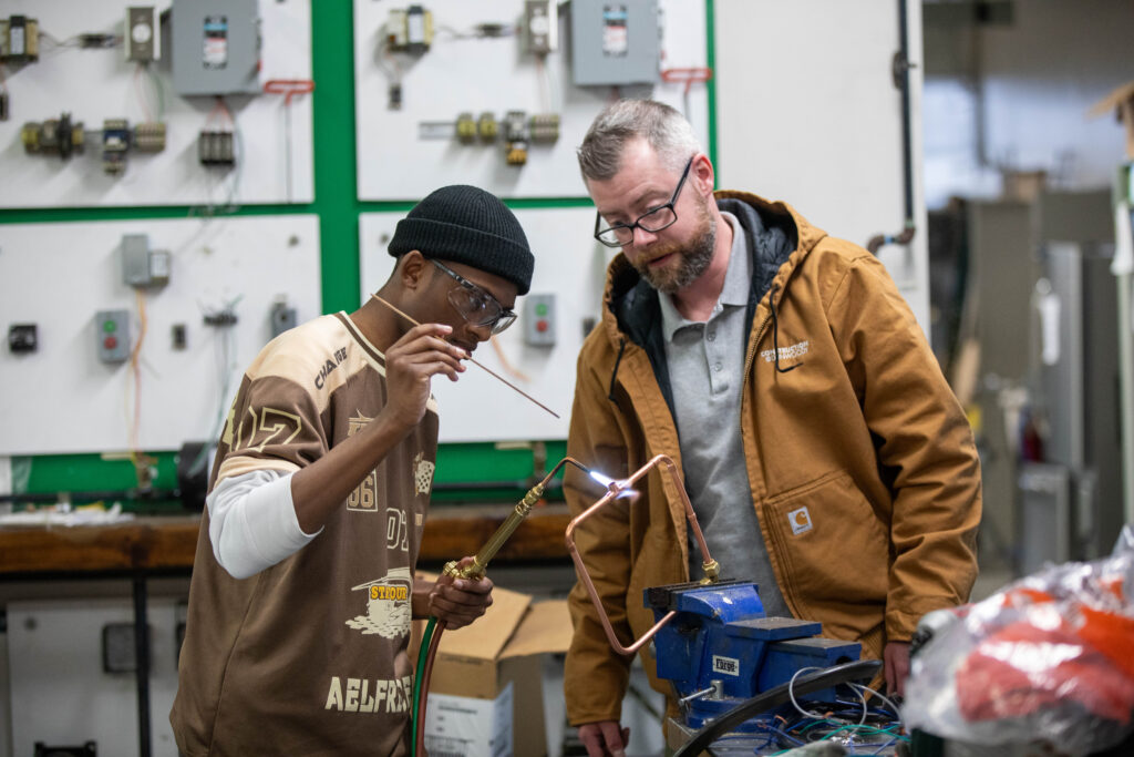 An HVAC faculty member reviews the technique of a student who is working on welding copper tubing
