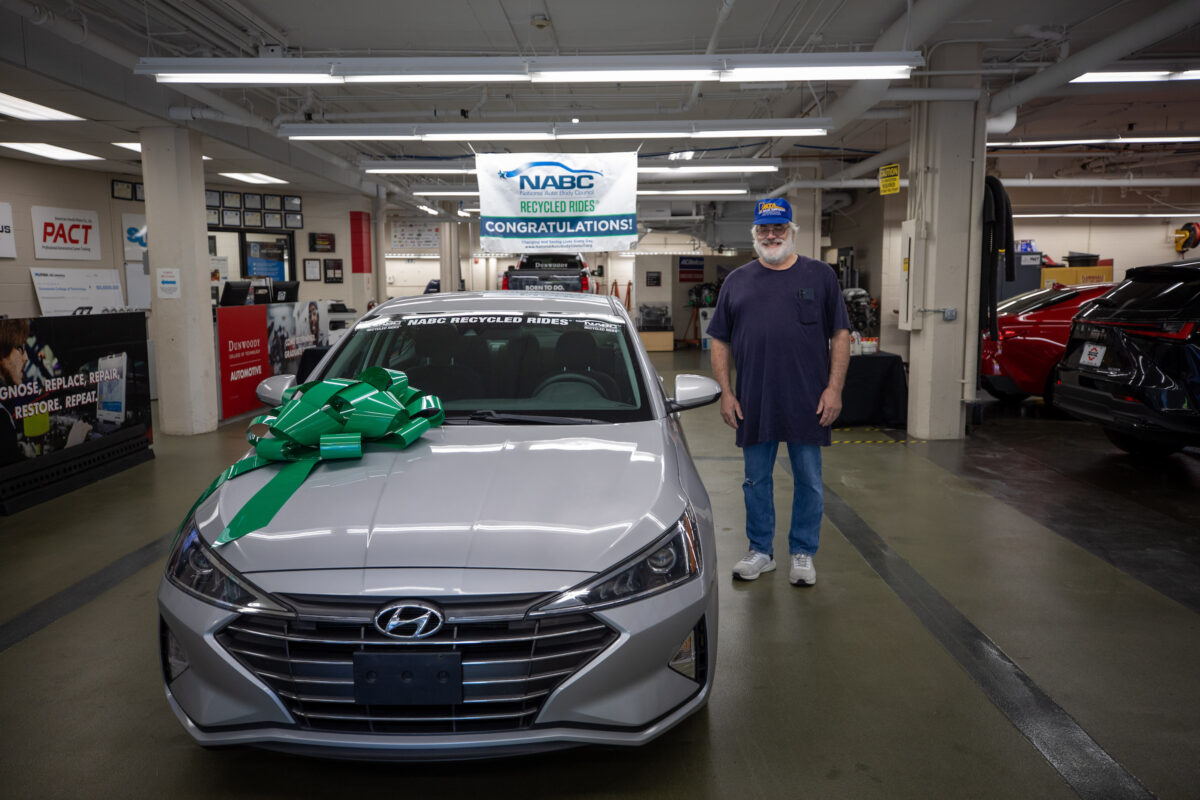 A person stands beside a silver Hyundai sedan adorned with a large green bow, under a banner celebrating NAABC Recycled Rides.