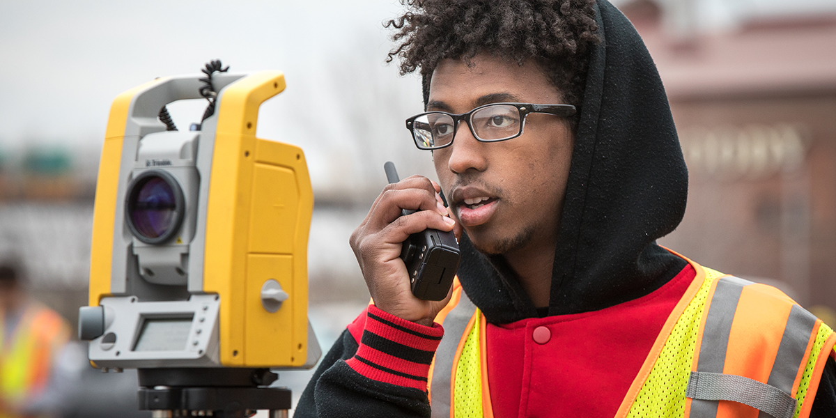 A student in the associates in civil engineering program standing next to a piece of surveying equipment and talking on walkie talkie to a fellow civil engineering technology student