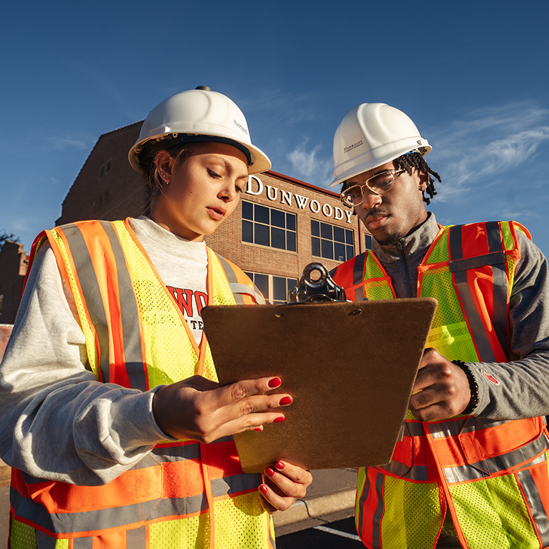 Two students in the Construction Project Management program stand outside wearing hard hats and holding a clip board.