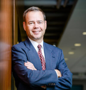 Photo of David Mortenson wearing a blue suit and dark red tie, standing with his arms crossed